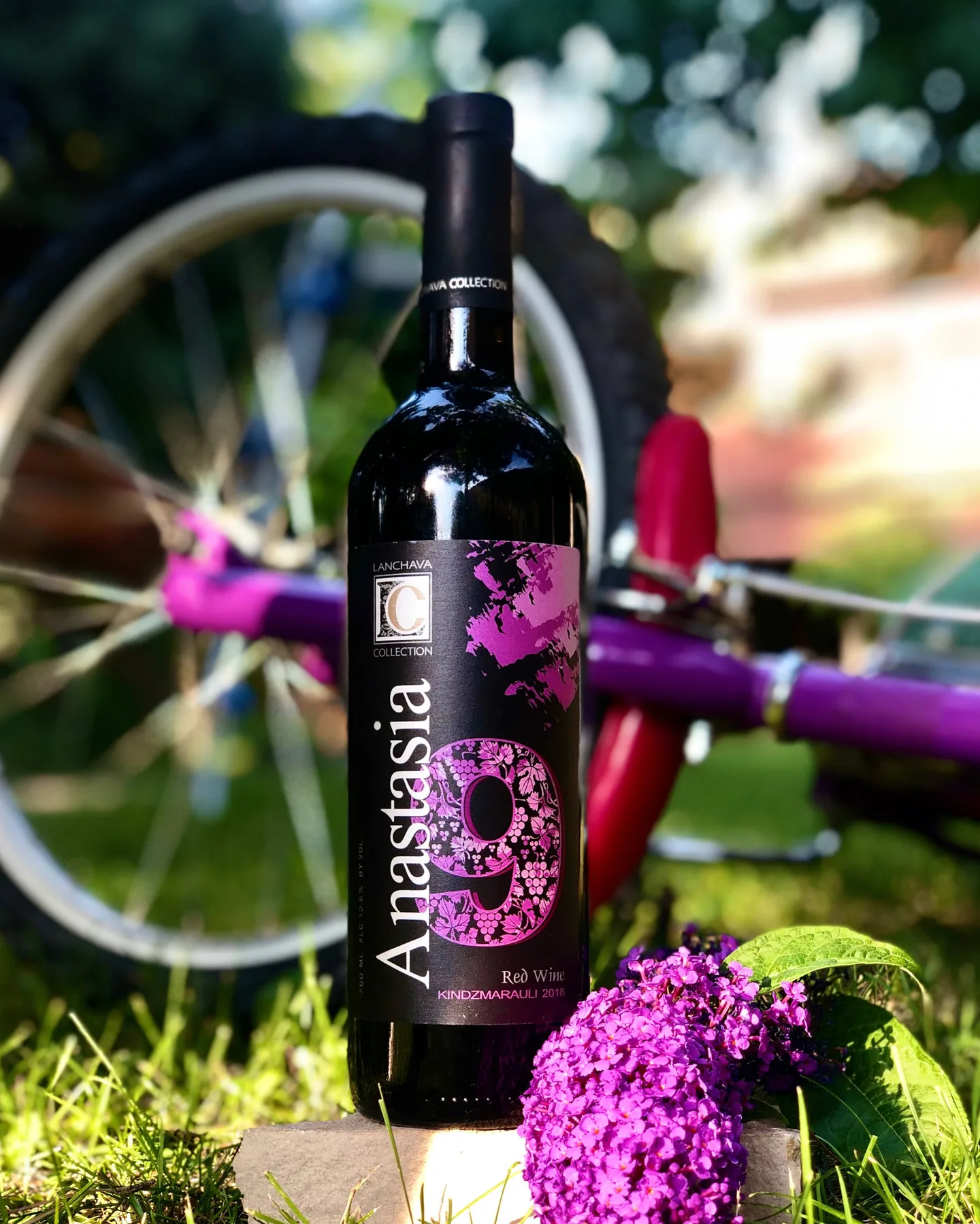 A bottle of Anastasia red wine standing in the grass beside a bicycle wheel and purple flowers