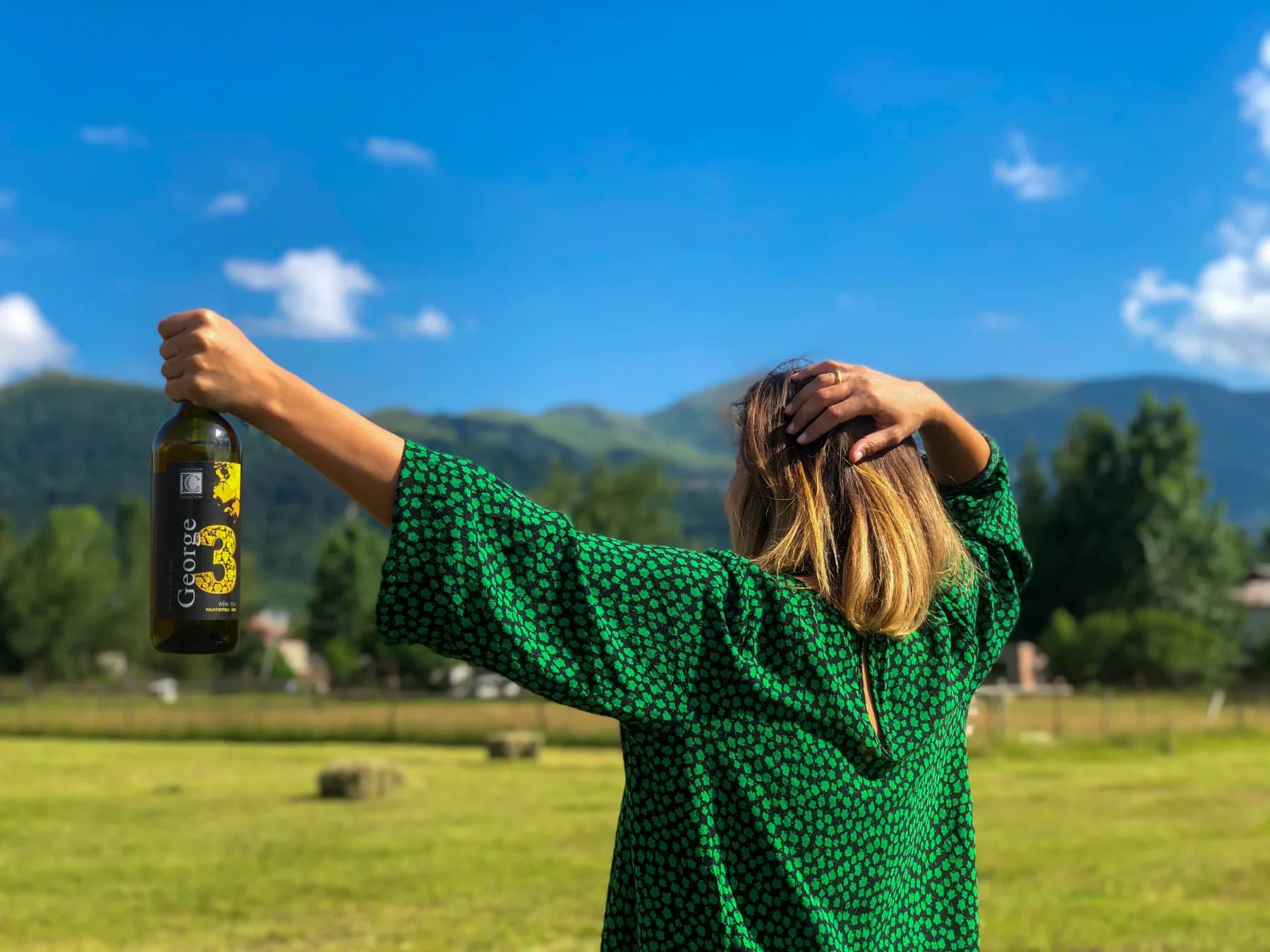 A person in a green dress raising a bottle of George white wine in a sunny meadow below green mountains